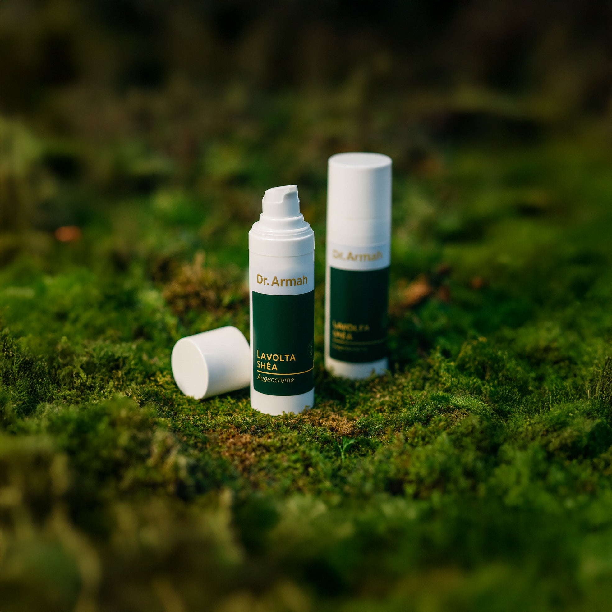 Ein stimmungsvolles Produktfoto zeigt zwei Flaschen "Dr. Armah LAVOLTA SHÉA Augencreme", die in einer natürlichen Umgebung auf einem dichten, grünen Moosbett arrangiert sind. Eine Flasche im Vordergrund ist geöffnet, während eine zweite, geschlossene Flasche im unscharfen Hintergrund steht.